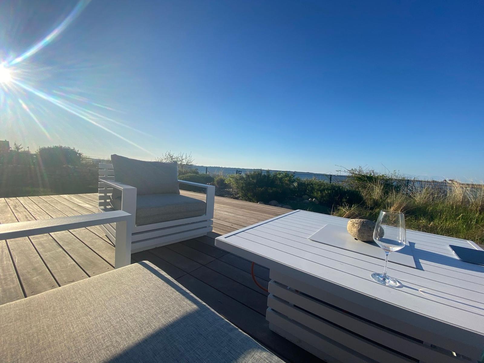 Terrasse mit weißem Tisch und Stuhl, Sonne, Blick auf Wasser und Gras.