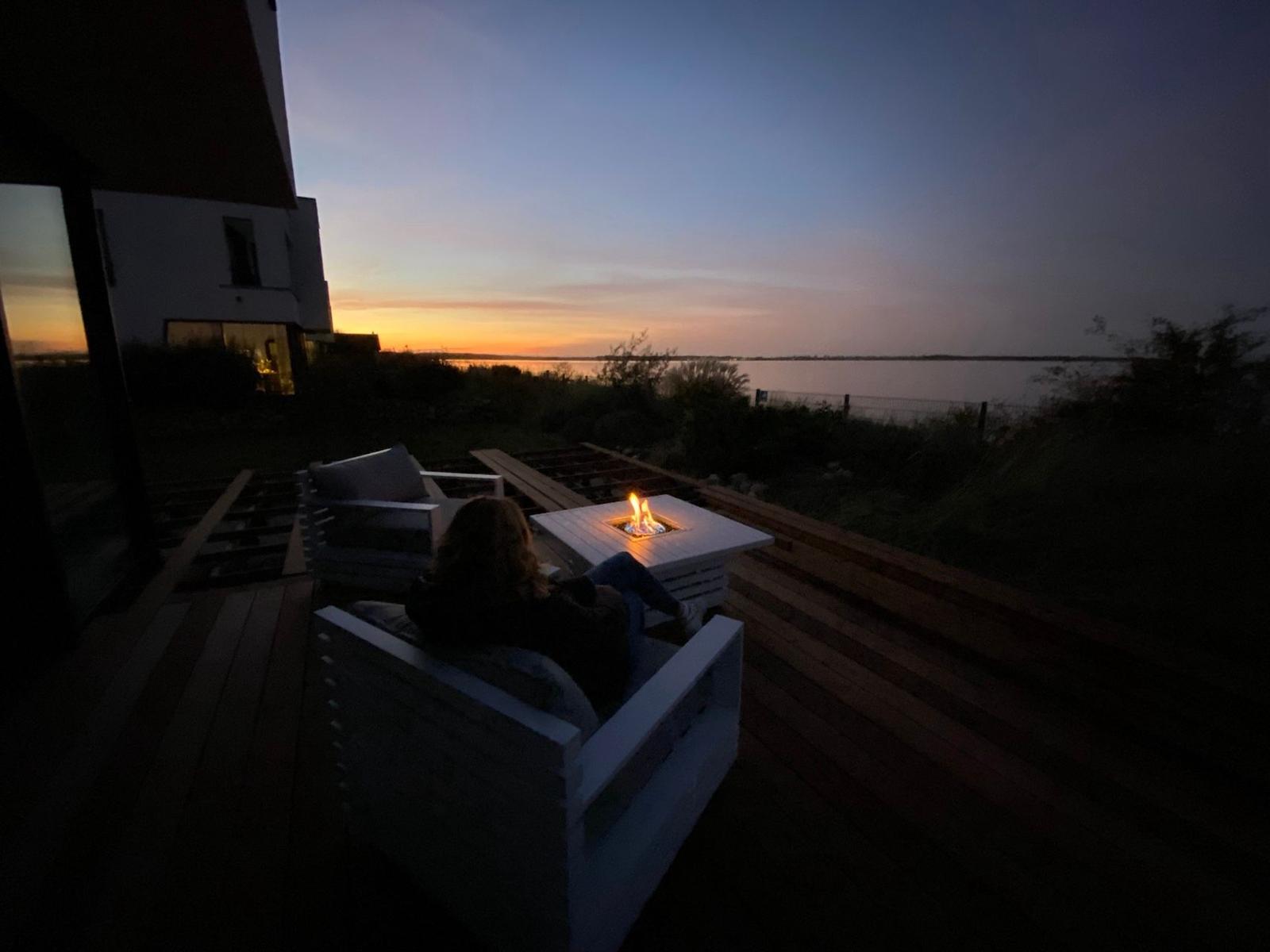 Person sitzt auf Terrasse mit Feuerkorb und Blick auf Wasser bei Sonnenuntergang.