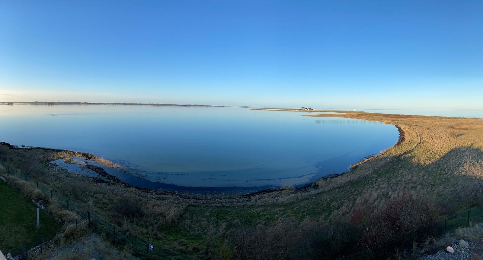 Panoramablick auf ruhiges Wasser und grasbewachsenen Uferbereich bei klarem Himmel.