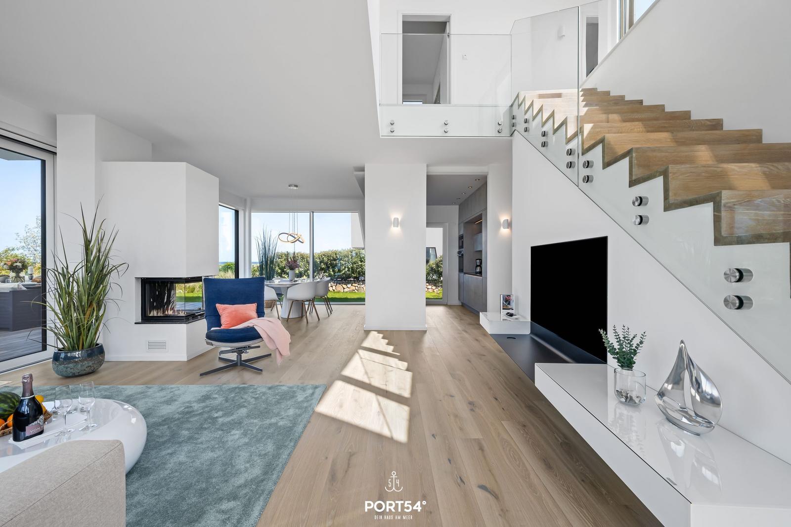 Wohn- und Essbereich mit Kamin, TV und Holztreppe. Durchgehendes Glasgeländer und großflächige Fenster.
