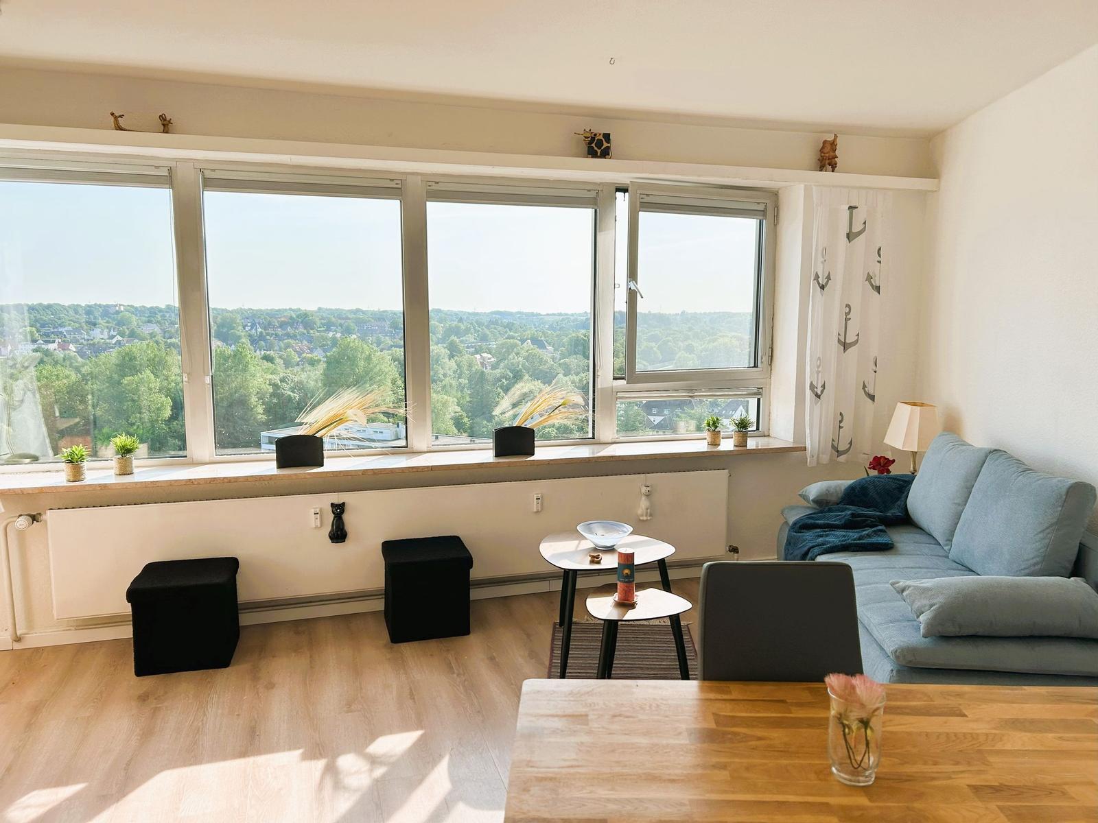 Wohnzimmer mit großem Fenster und Blick auf die Stadt. Sofa, Tische und Holzboden.