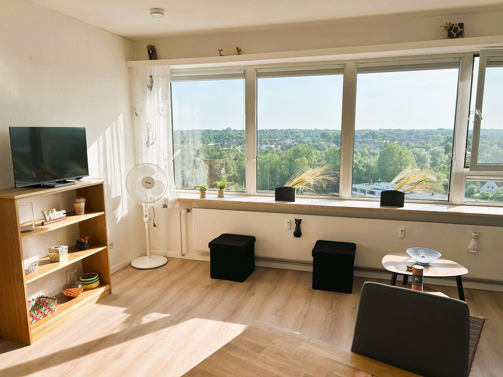 Wohnzimmer mit großem Fenster, TV und Sitzgelegenheiten.