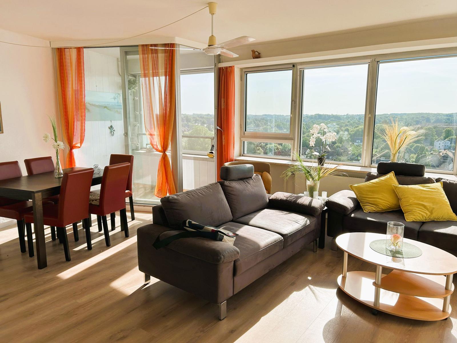 Wohn- und Essbereich mit Sofa, Tisch und großen Fenstern. Blick auf grüne Landschaft.