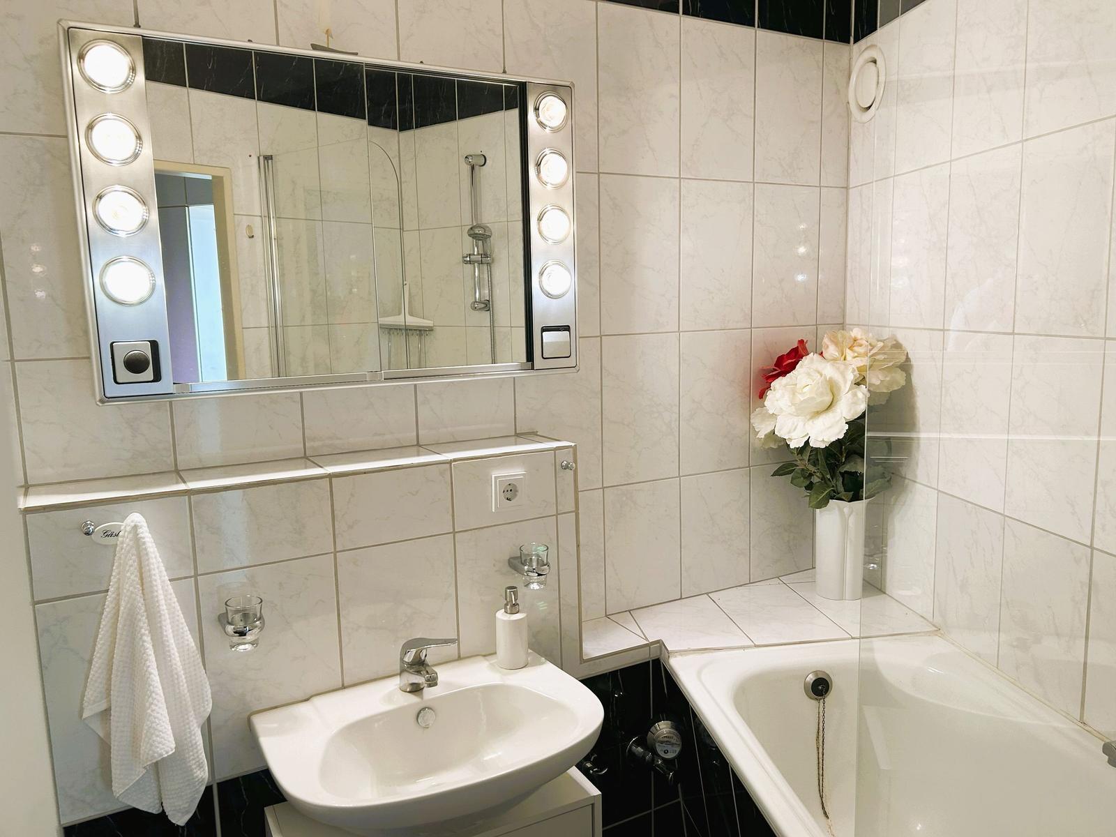 Badezimmer mit Waschbecken, Dusche und Badewanne. Spiegel mit Beleuchtung und Blumen auf dem Rand.