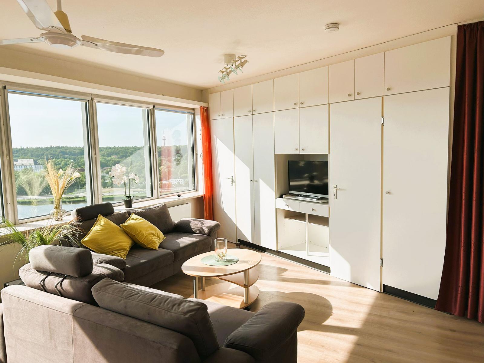 Wohnzimmer mit Sofa, Couchtisch und TV-Einbauwand. Große Fenster mit Blick auf Wasser.