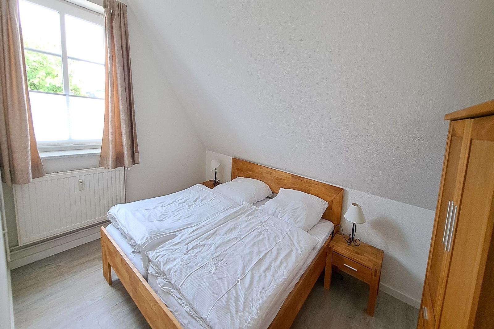 Schlafzimmer mit Doppelbett, Nachttisch und Kleiderschrank. Fenster mit Vorhängen.