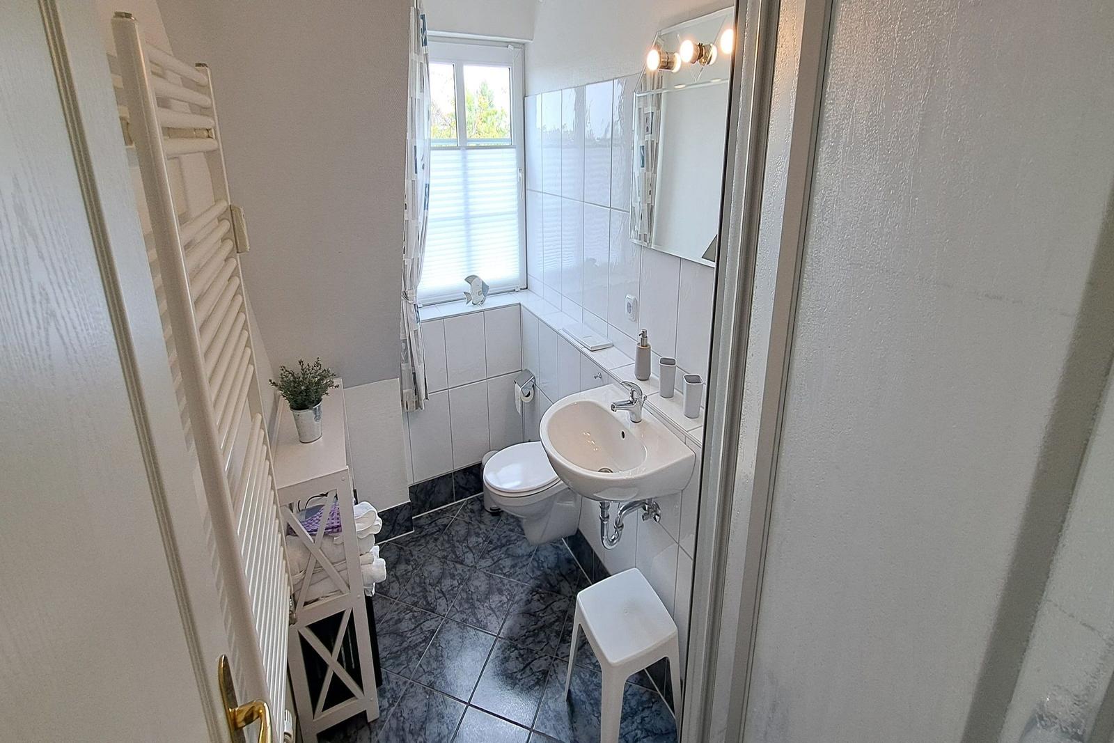 Badezimmer mit Toilette, Waschbecken und Fenster. Weiße Fliesen und Duschwanne.