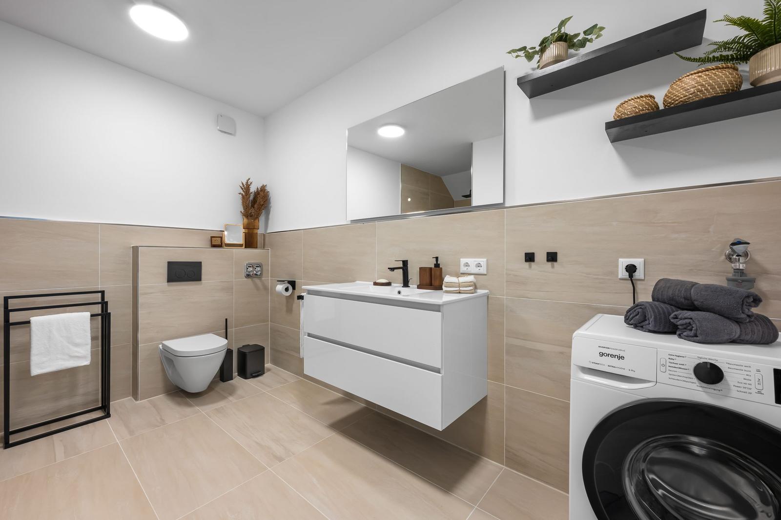 Badezimmer mit Waschmaschine, Spiegel und Toilette. Wandfliesen und Holzakzente.