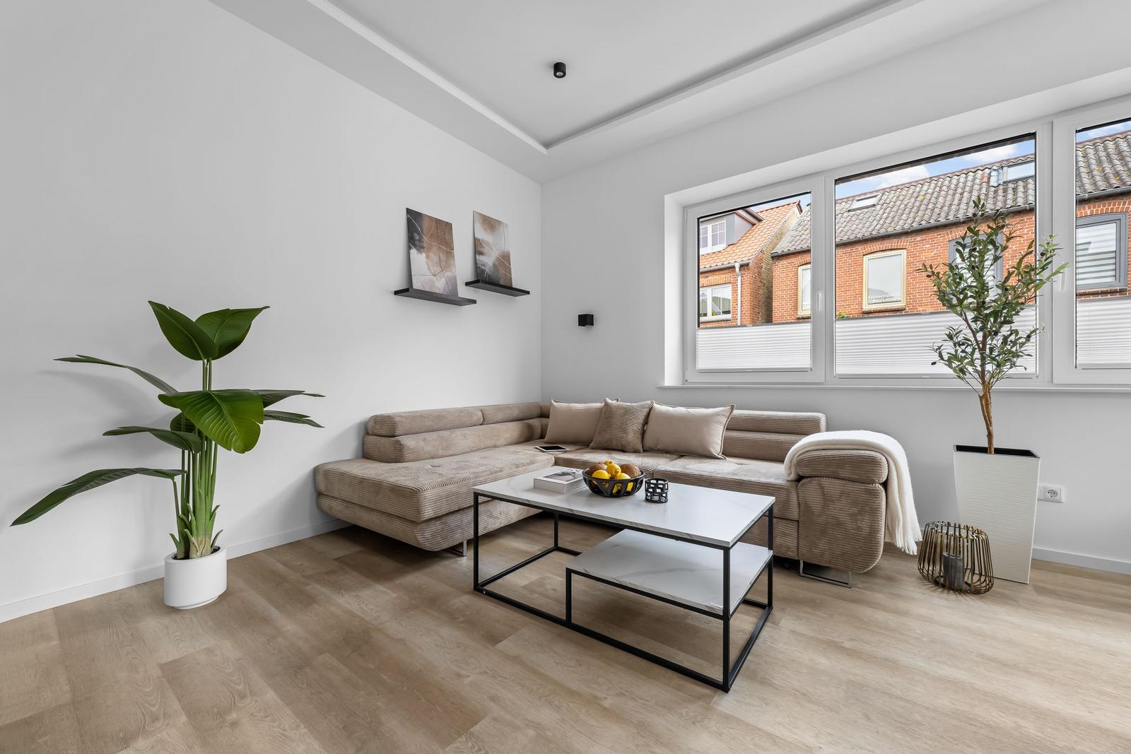 Wohnzimmer mit Ecksofa, Tisch und Pflanzen. Fenster zeigen auf Straßenansicht.