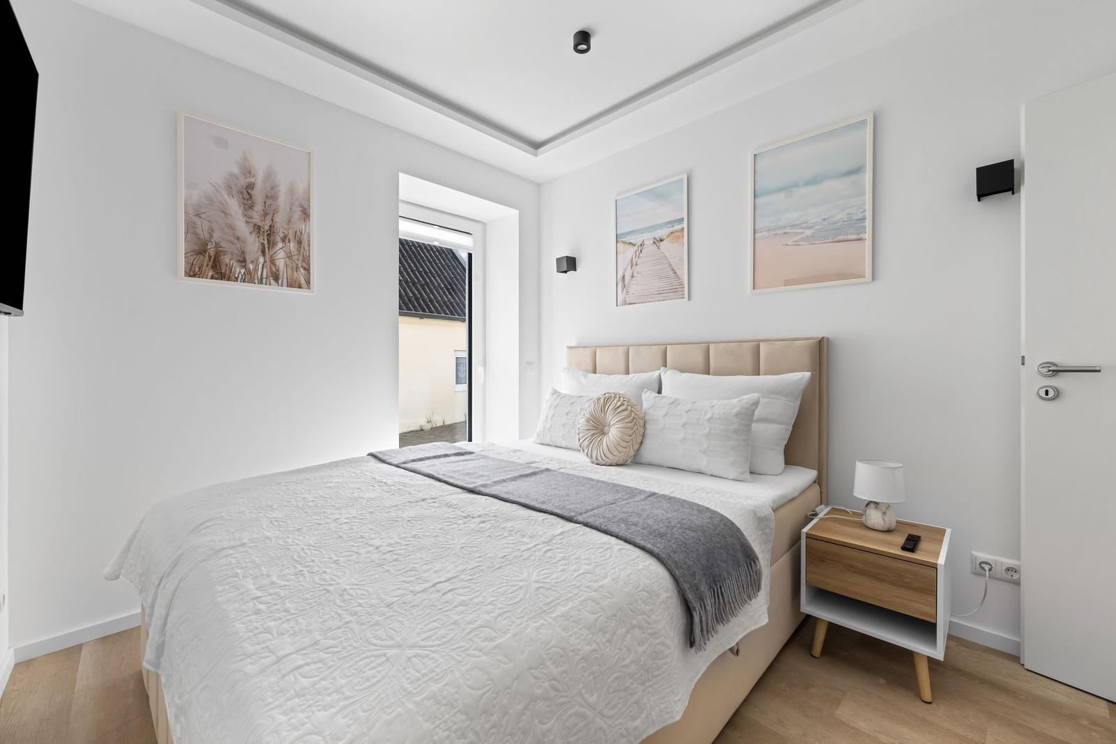 Schlafzimmer mit Bett, Nachttisch und Wandbildern. Tür führt nach draußen.