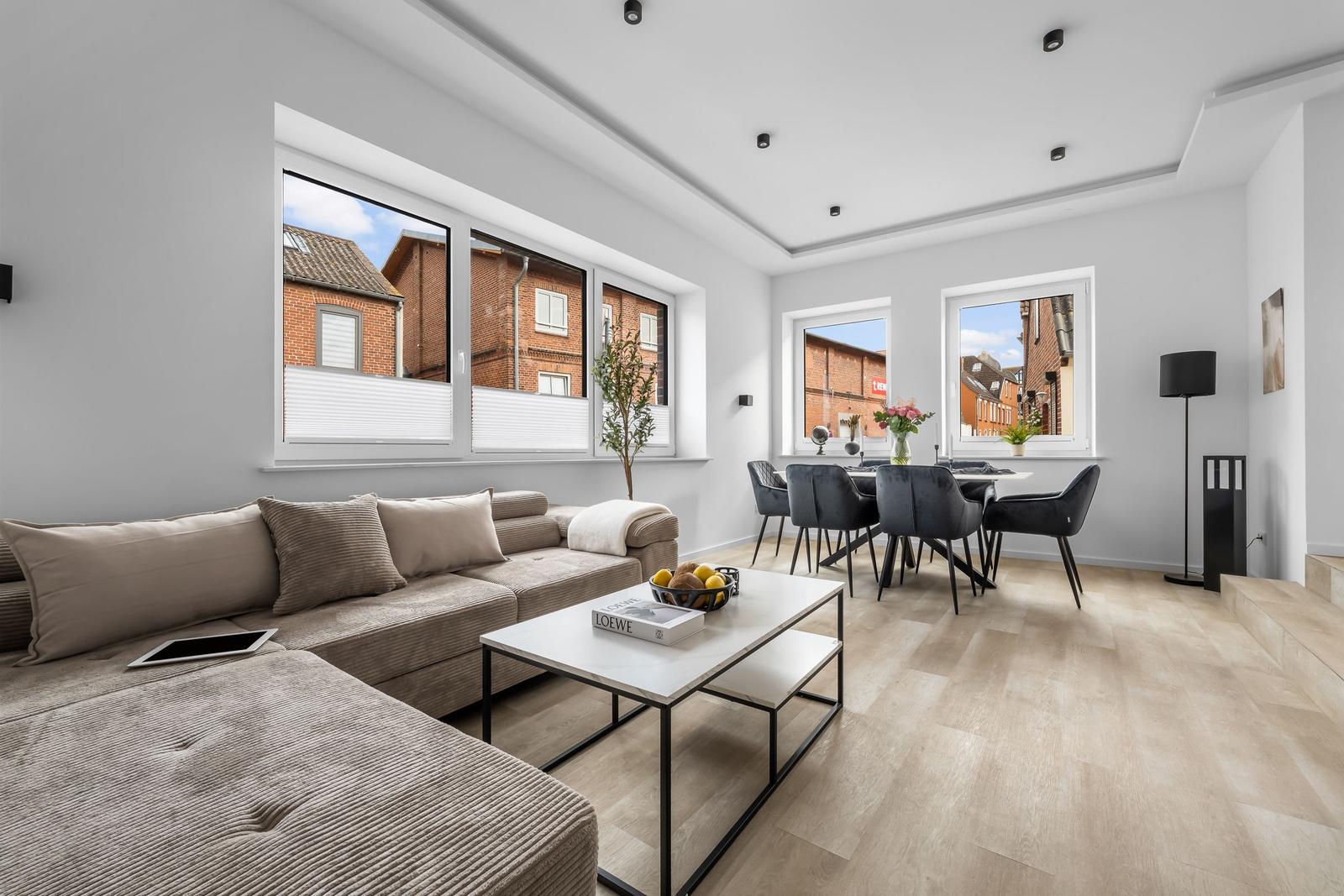 Wohn- und Essbereich mit Sofa, Tisch und Stühlen. Große Fenster mit Blick auf Gebäude.