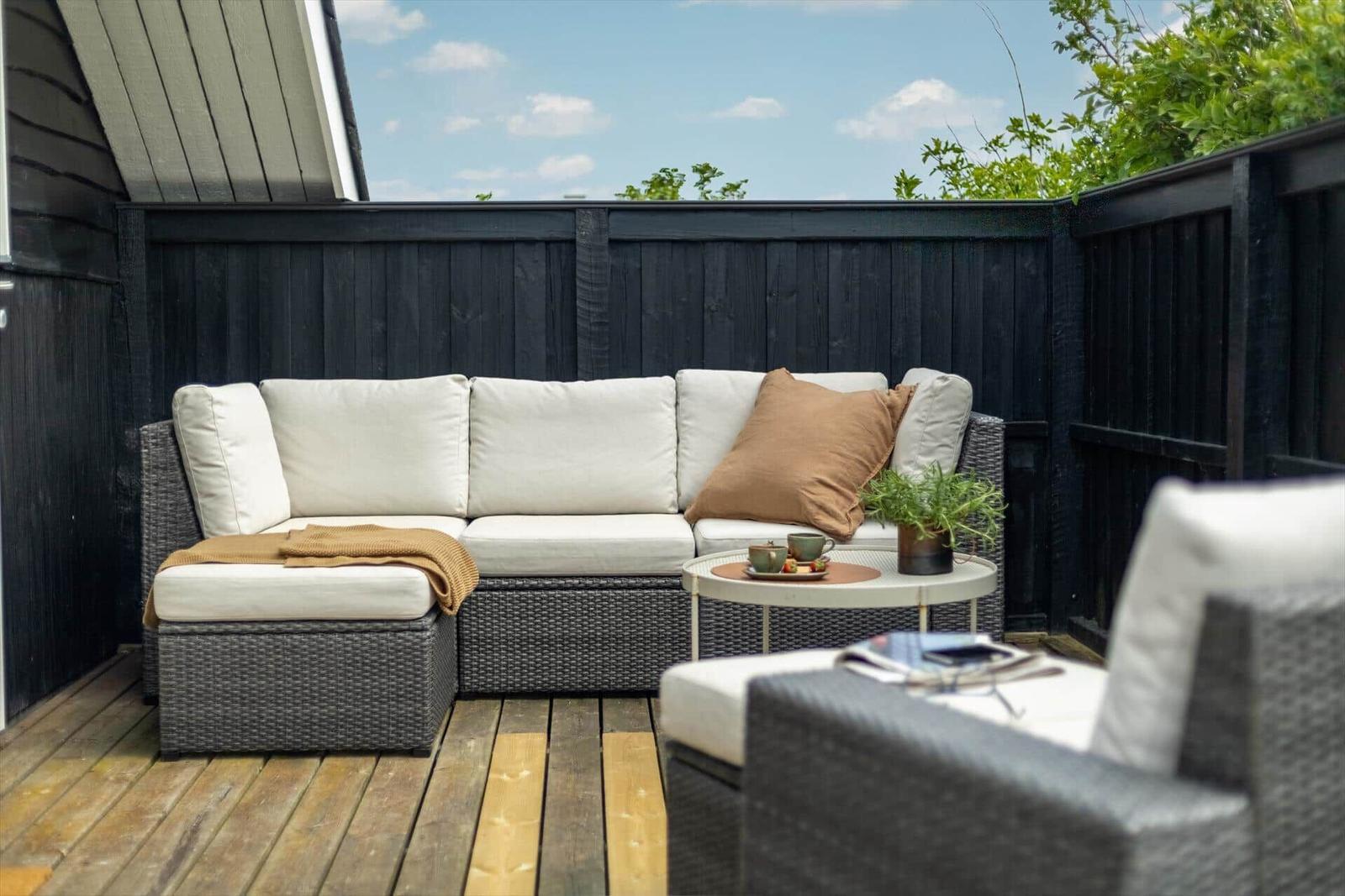 Terrasse mit Rattanmöbeln, Kissen und Tisch. Holzdecke und schwarzer Zaun.