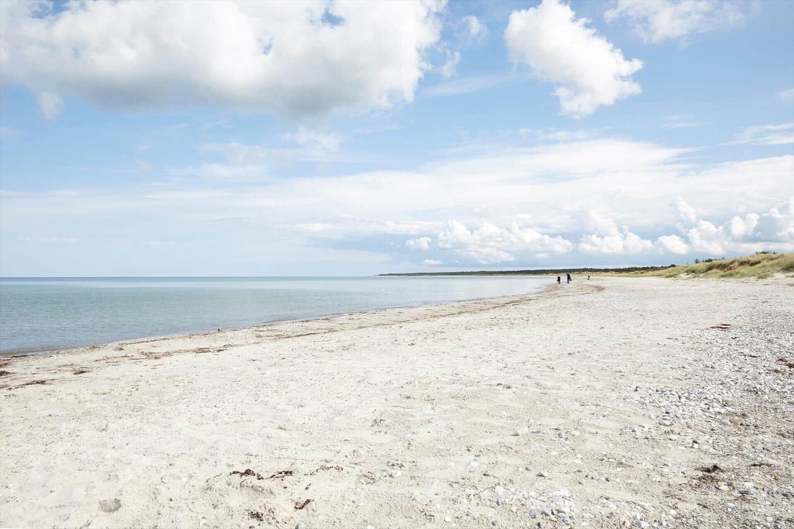 Weiterer Strandabschnitt mit weißen Sand und ruhigem Meer unter blauem Himmel mit weißen Wolken.