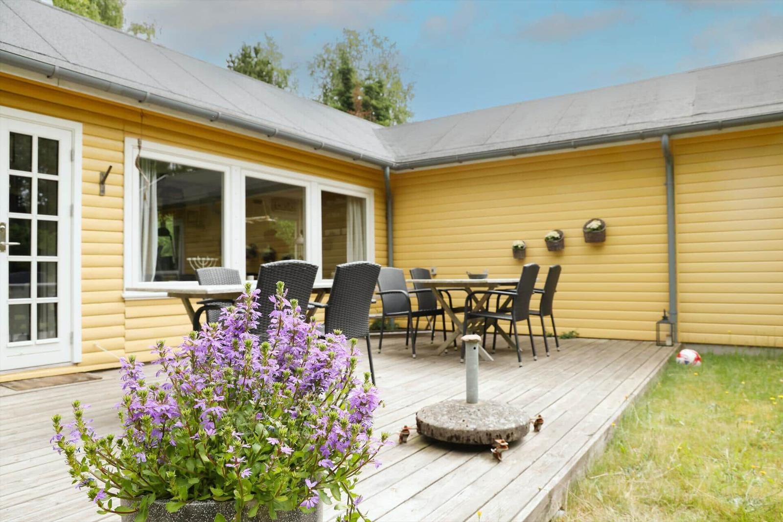 Helle gelbe Holzhausfassade mit Terrasse, Tisch, Stühlen und Blumen.
