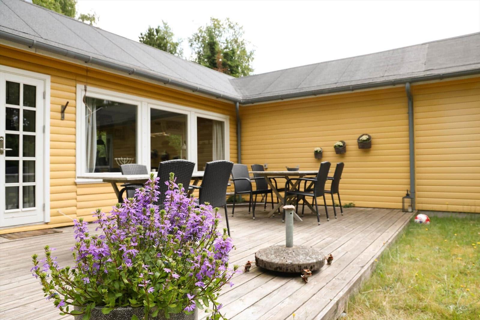 Holzterrasse mit Tisch, Stühlen und Blumenbeet vor gelbem Haus.