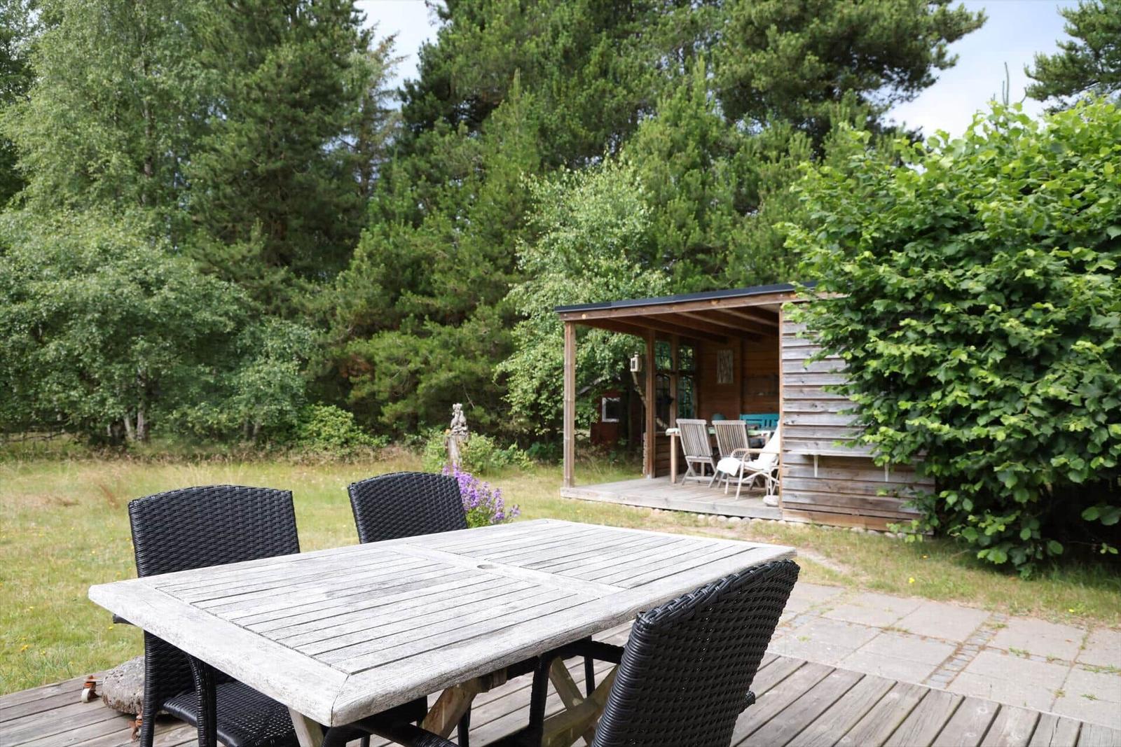 Holzterrasse mit Tisch und Stühlen, umgeben von Grün und einem Holzhaus.