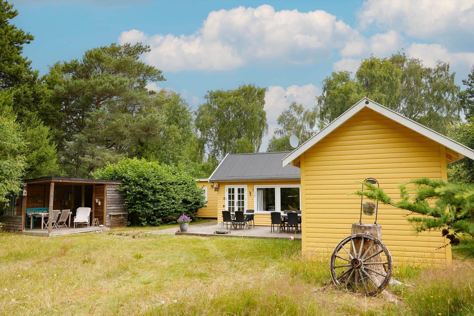 Gelbes Ferienhaus mit Terrasse, Garten und Holzständer. Hintergrund: Bäume und blauer Himmel.