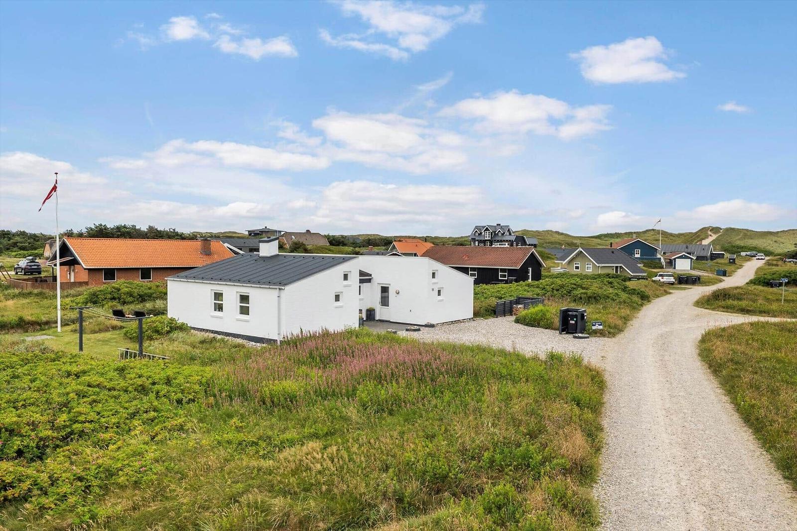 Weißes Haus mit dunklem Dach, umgeben von Gras und Dünen. Straße führt zu weiteren Häusern.