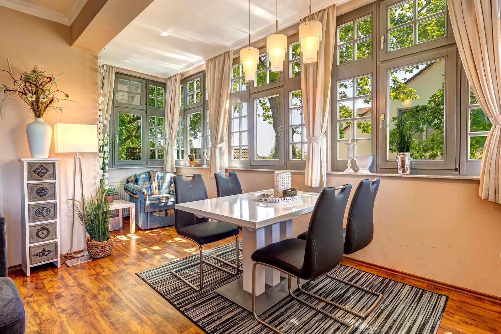 Esszimmer mit Tisch, Stühlen und großen Fenstern. Holzboden und Vorhänge.