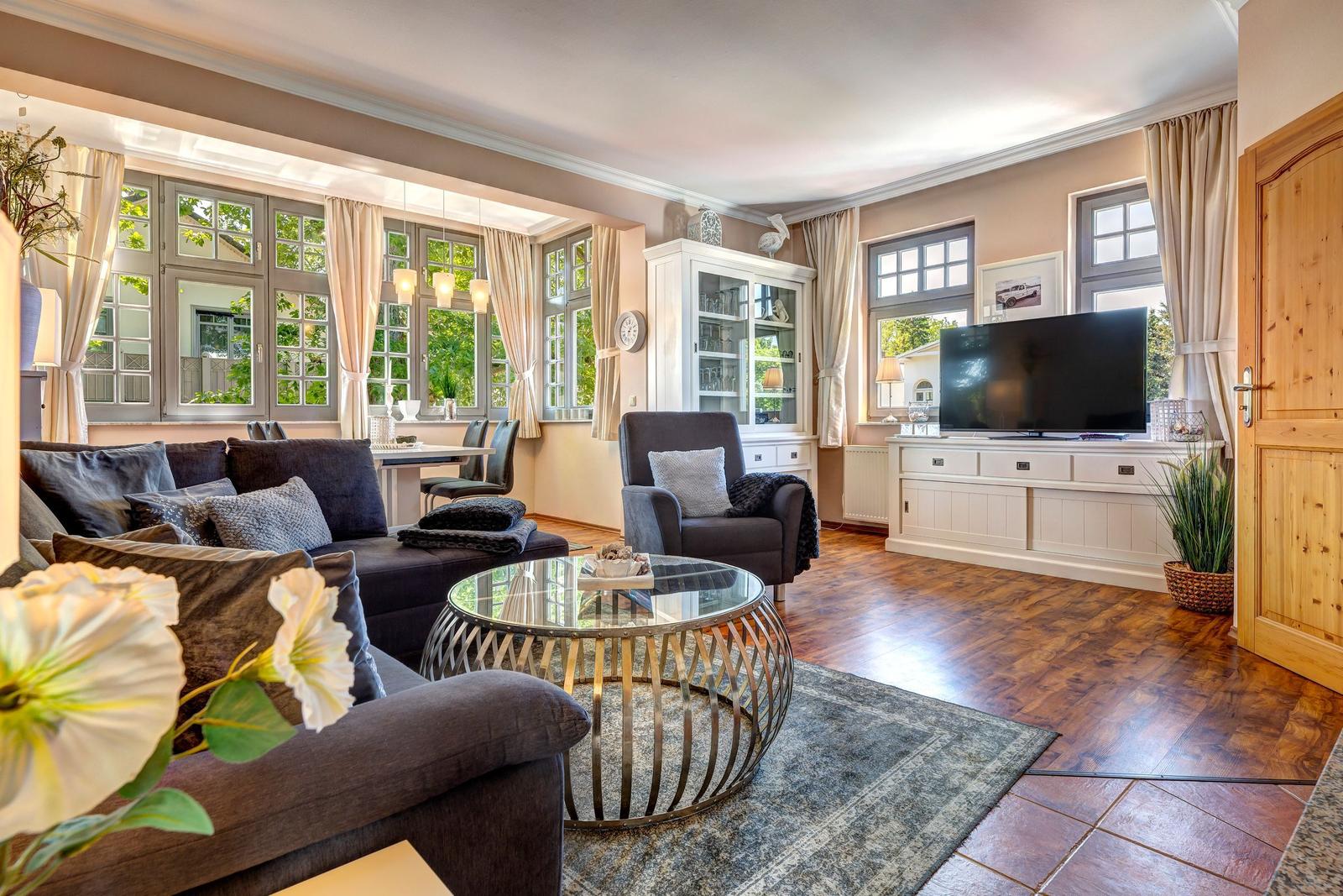 Wohnzimmer mit Sofa, Sessel, Glas-Tisch und TV. Holzboden und große Fenster.