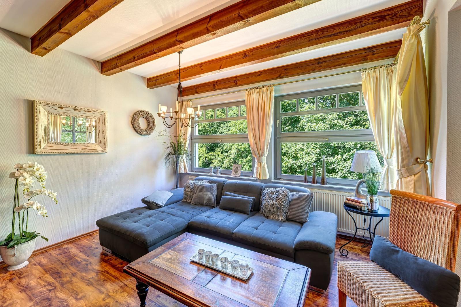 Ein Wohnzimmer mit grauem Sofa, Holzbalken und großem Fenster mit Blick auf Grün.