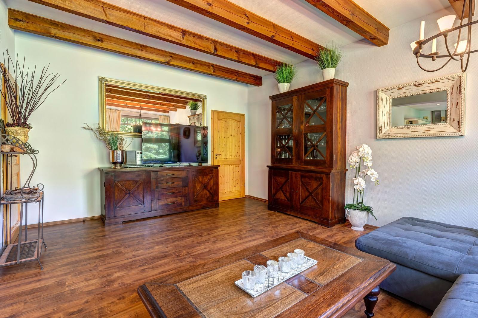 Wohnzimmer mit Holzboden, TV-Schrank und Glaswand-Schrank. Deckenbalken und Blumen.