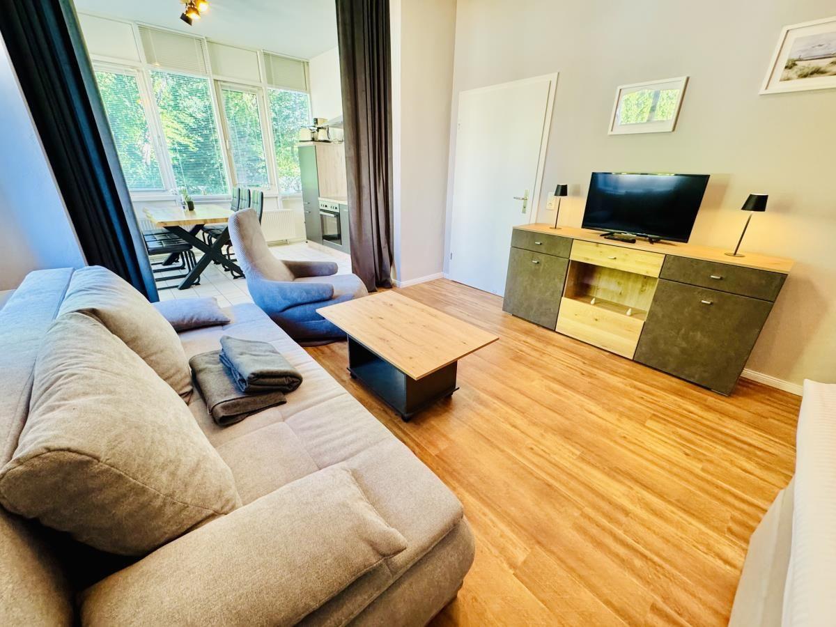 Wohnzimmer mit Sofa, Couchtisch und Fernseher. Durchgang zur Küche mit Tisch und Stühlen.