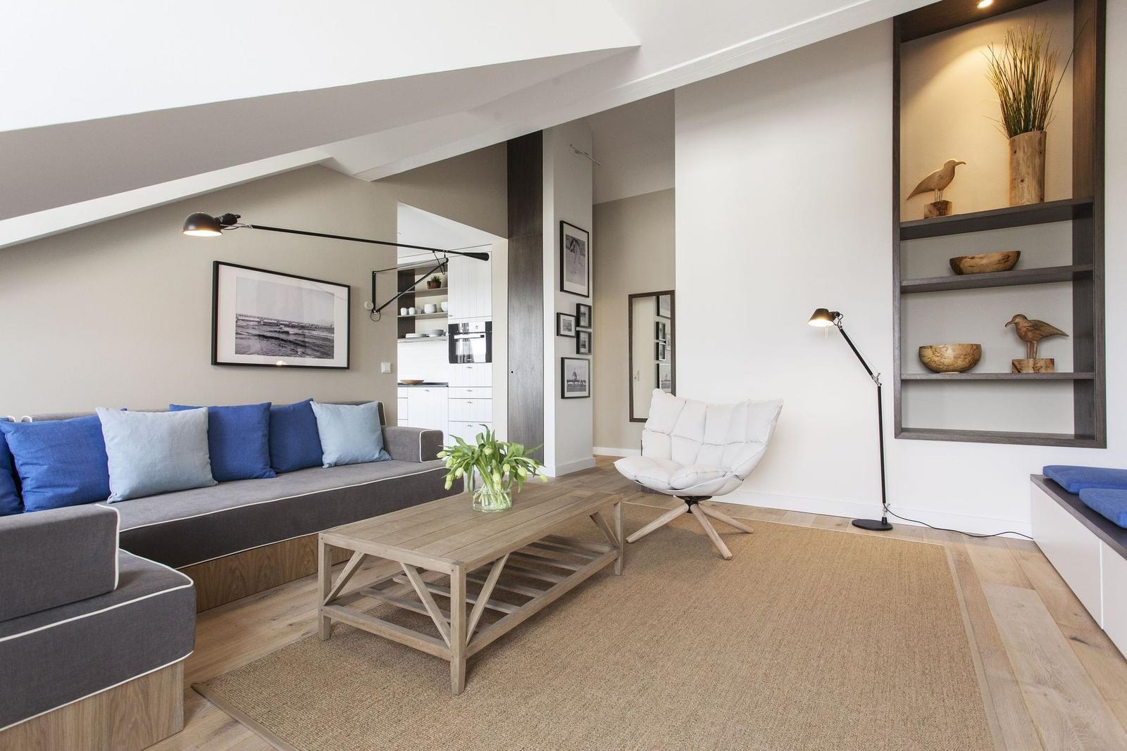 Wohnzimmer mit Sofa, Sessel und Holztisch. Regal mit Dekorationsgegenständen. Küchenbereich im Hintergrund.