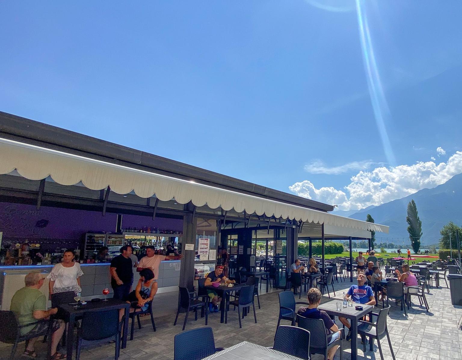 Außenbereich mit Tischen, Gästen und Bergblick unter blauem Himmel.