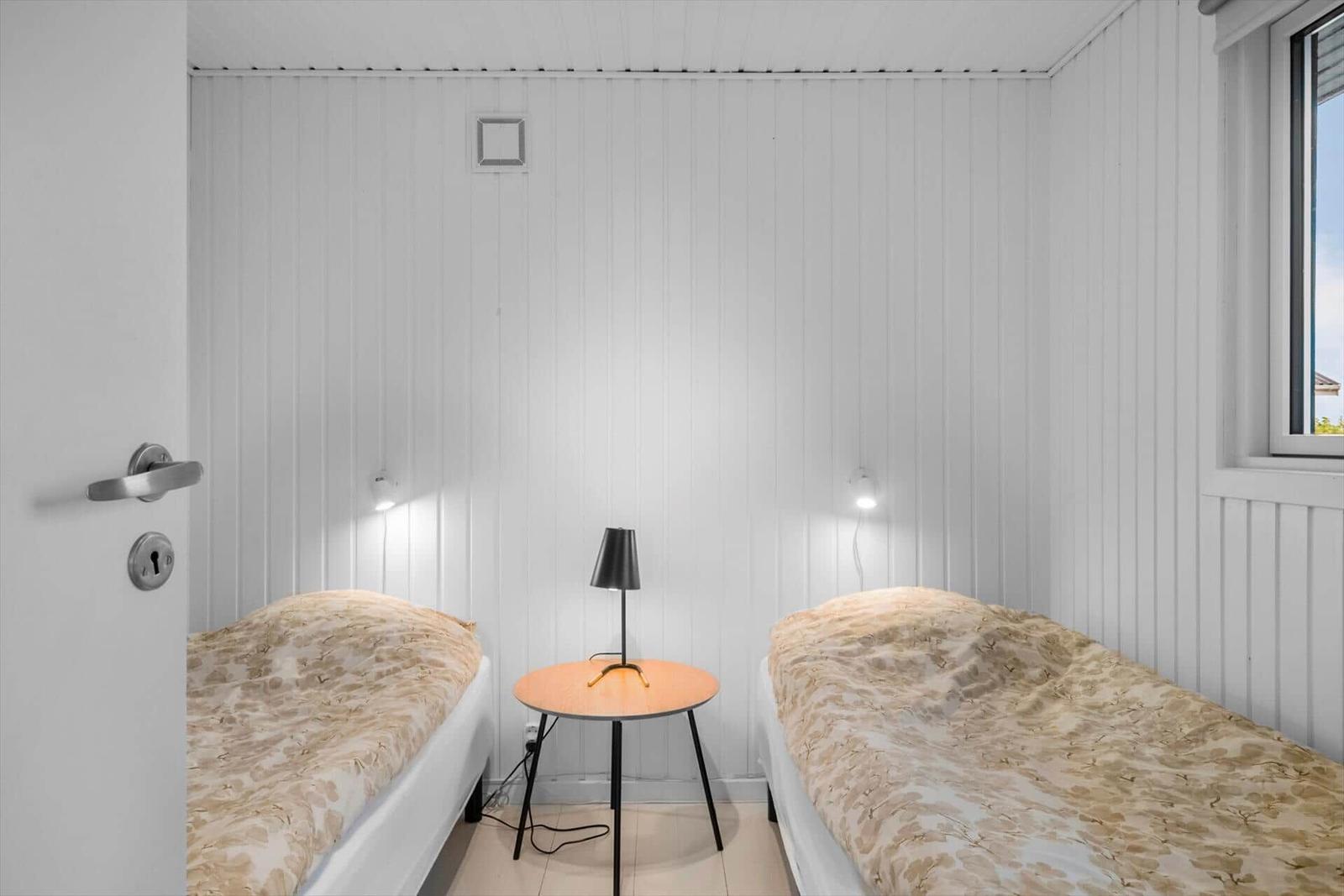 Zwei Einzelbetten mit Tisch und Lampe in einem Raum mit weißen Wänden.