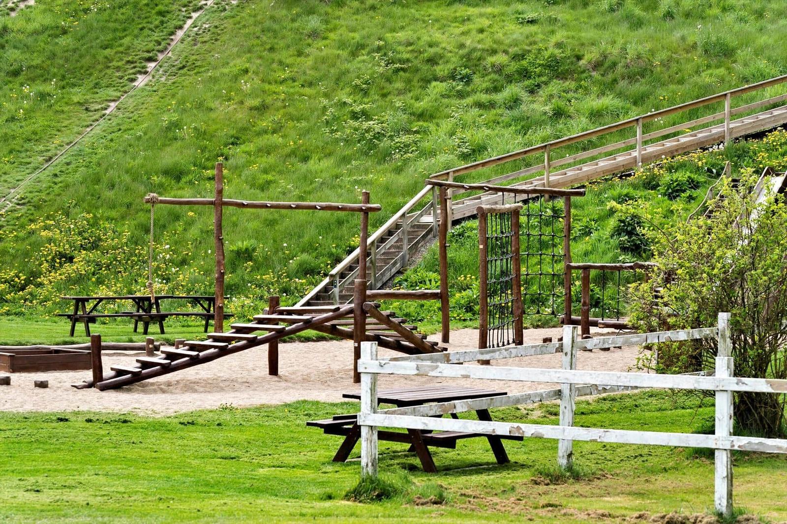 Holzspielplatz mit Rutsche, Klettergerüst und Picknicktischen auf grünem Hang.