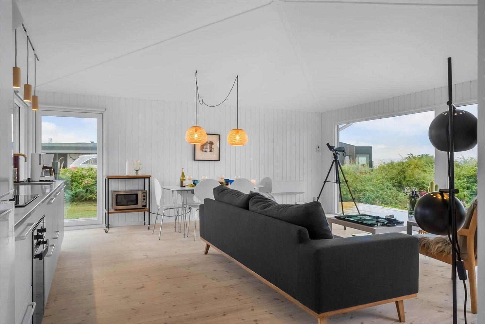 Wohnbereich mit Sofa, Esstisch und Küchenzeile. Große Fenster bieten Blick auf die Umgebung.