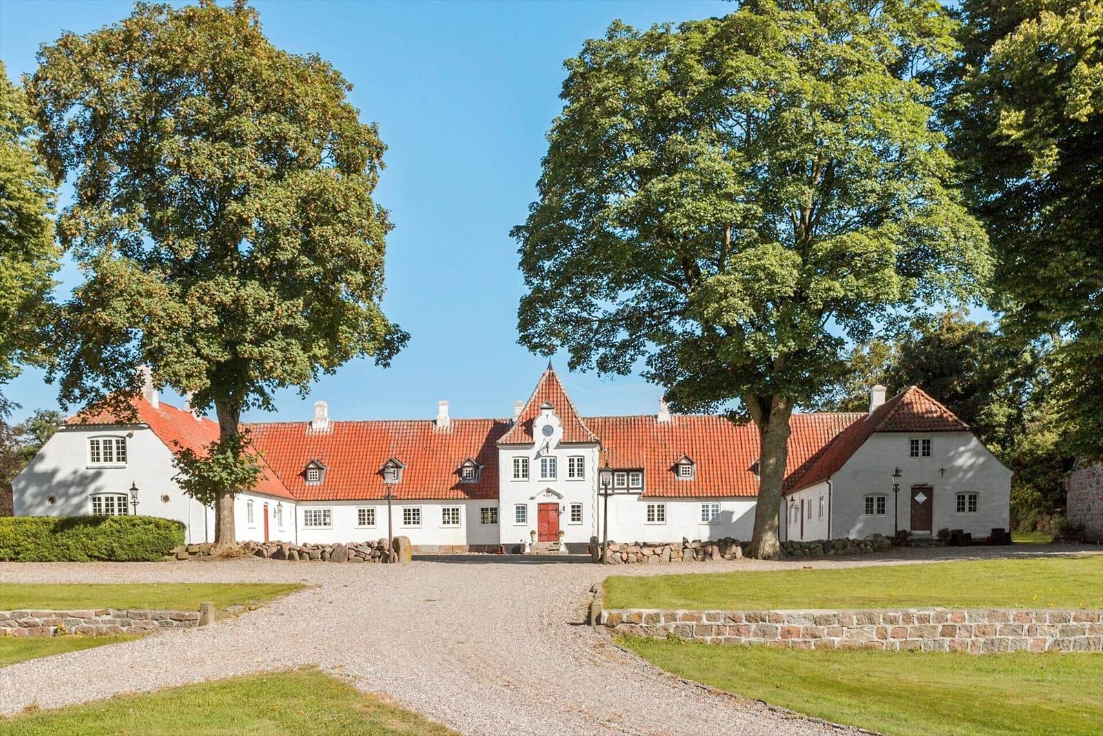 White villa with red roof, surrounded by trees and lawn.