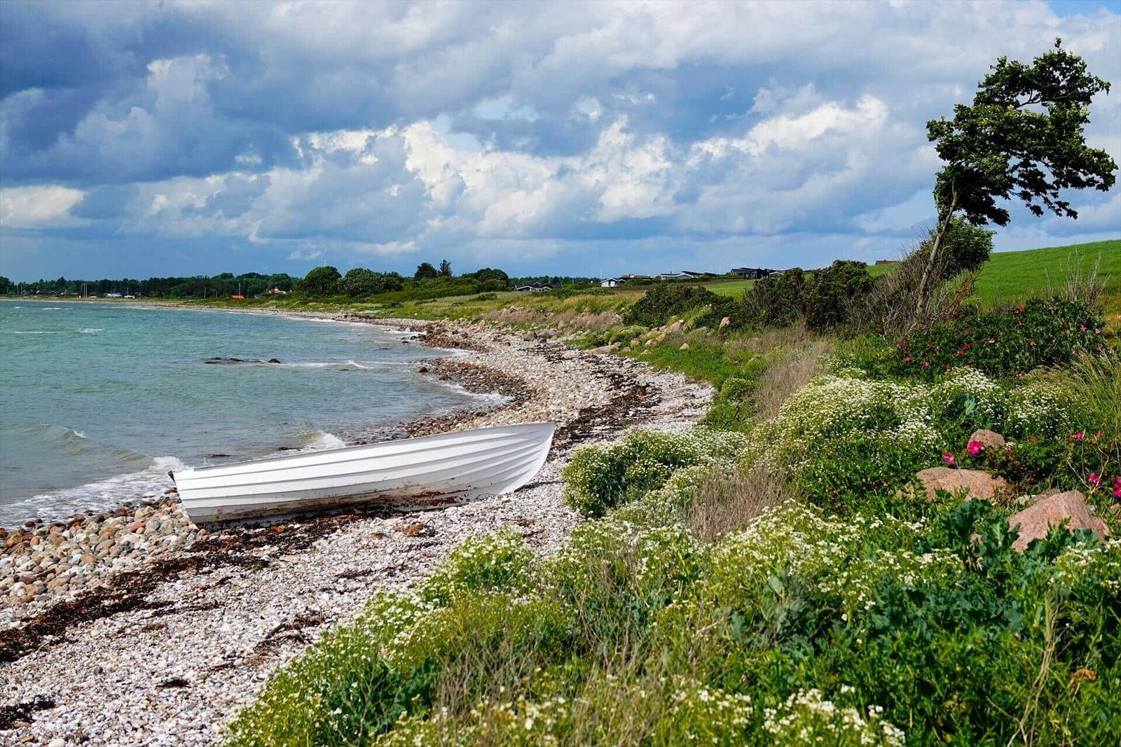 White boat rests on rocky shore. Greenery and flowers line the waterfront area.