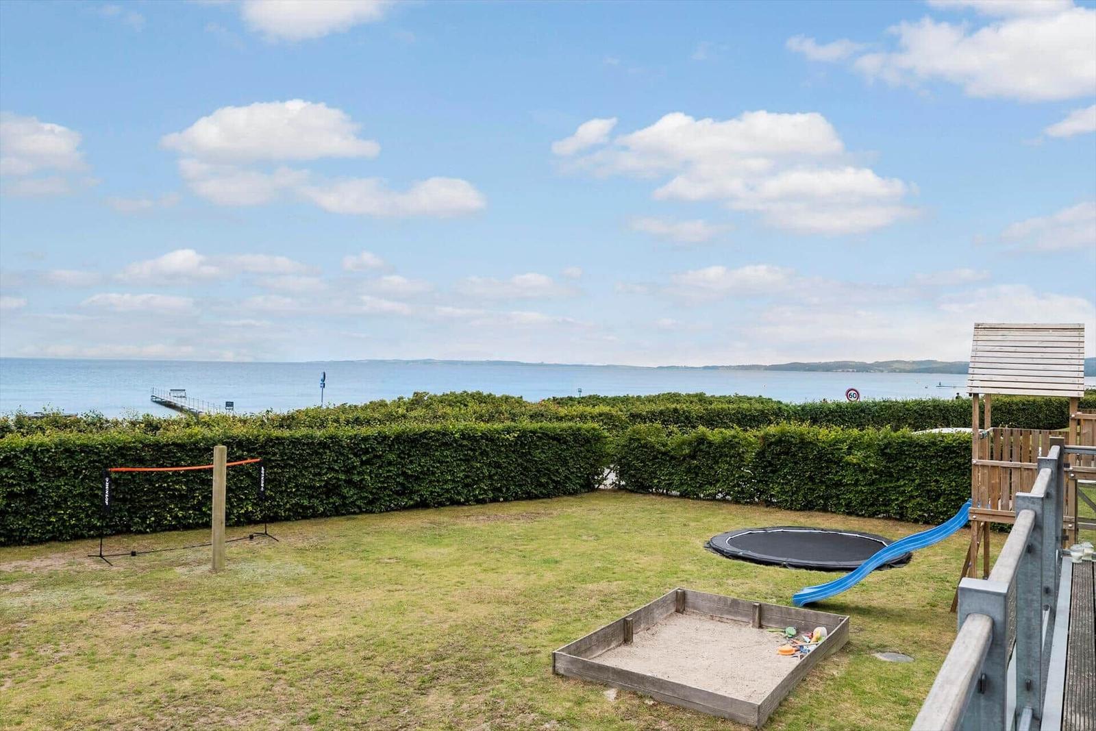 Grüner Garten mit Sandkasten, Trampolin und Spielplatz. Blick aufs Meer durch Hecke.