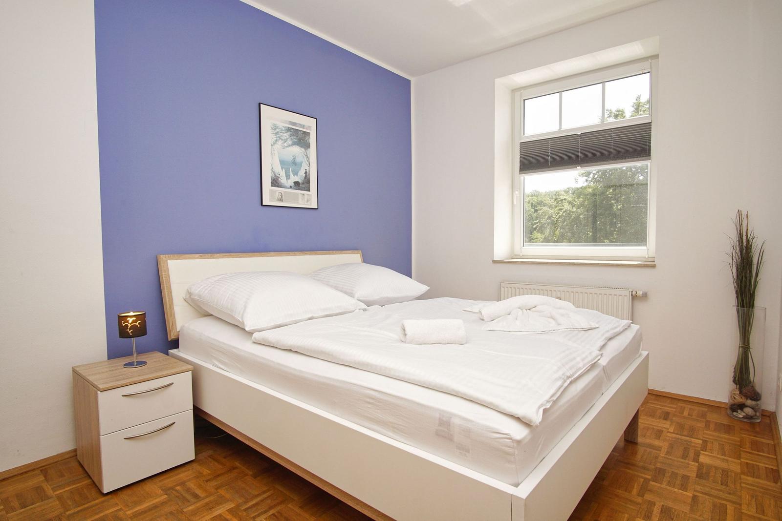 Schlafzimmer mit Bett, Nachttisch und Fenster. Wand in Blau. Holzfußboden.