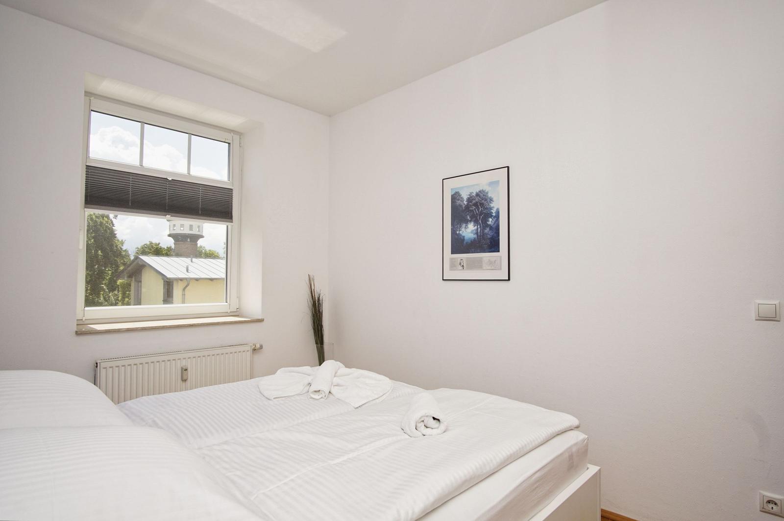 Ein Schlafzimmer mit weißem Bett, Fenster und Bild an der Wand.