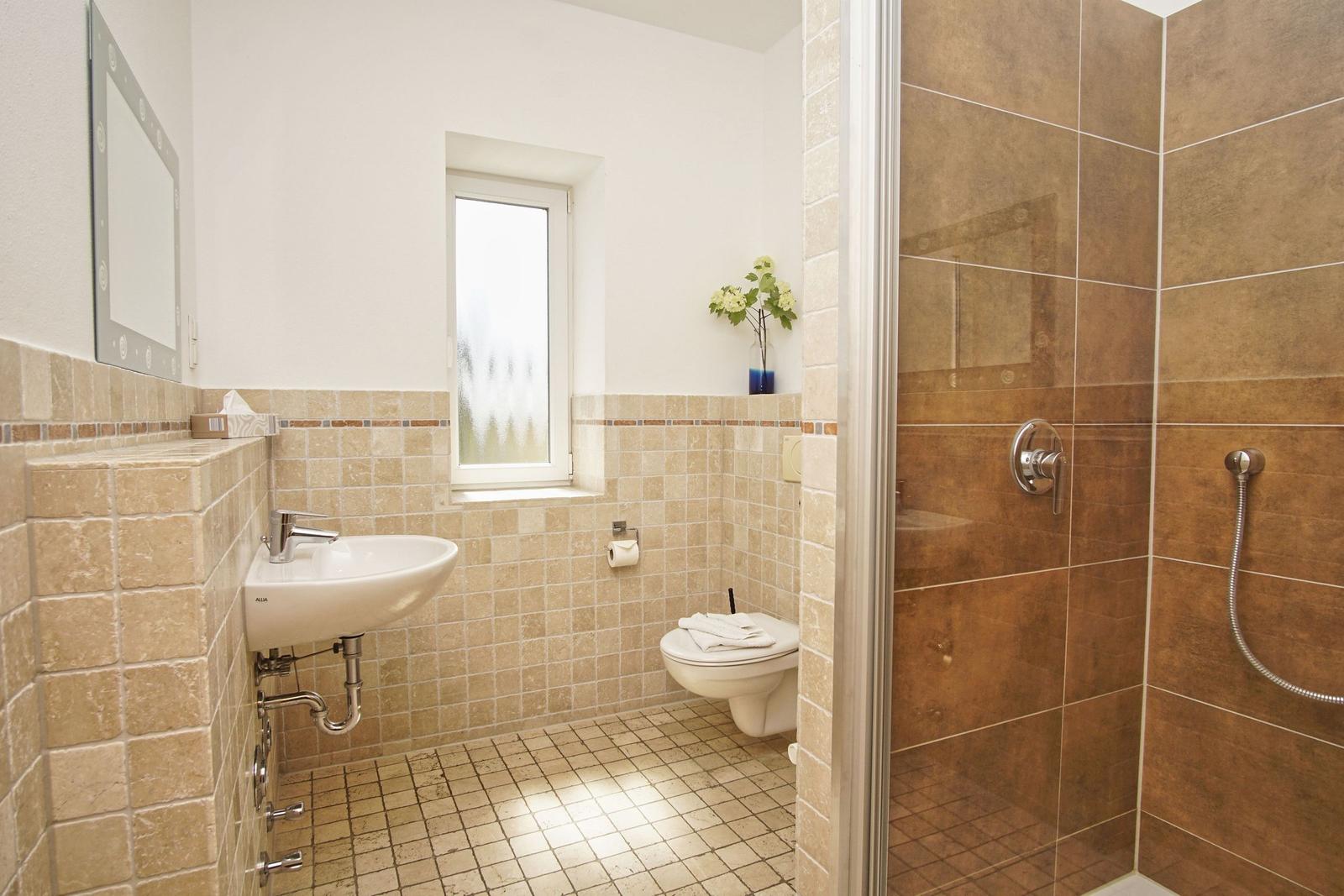 Badezimmer mit Dusche, Waschbecken und Toilette. Fliesen und Fenster sichtbar.