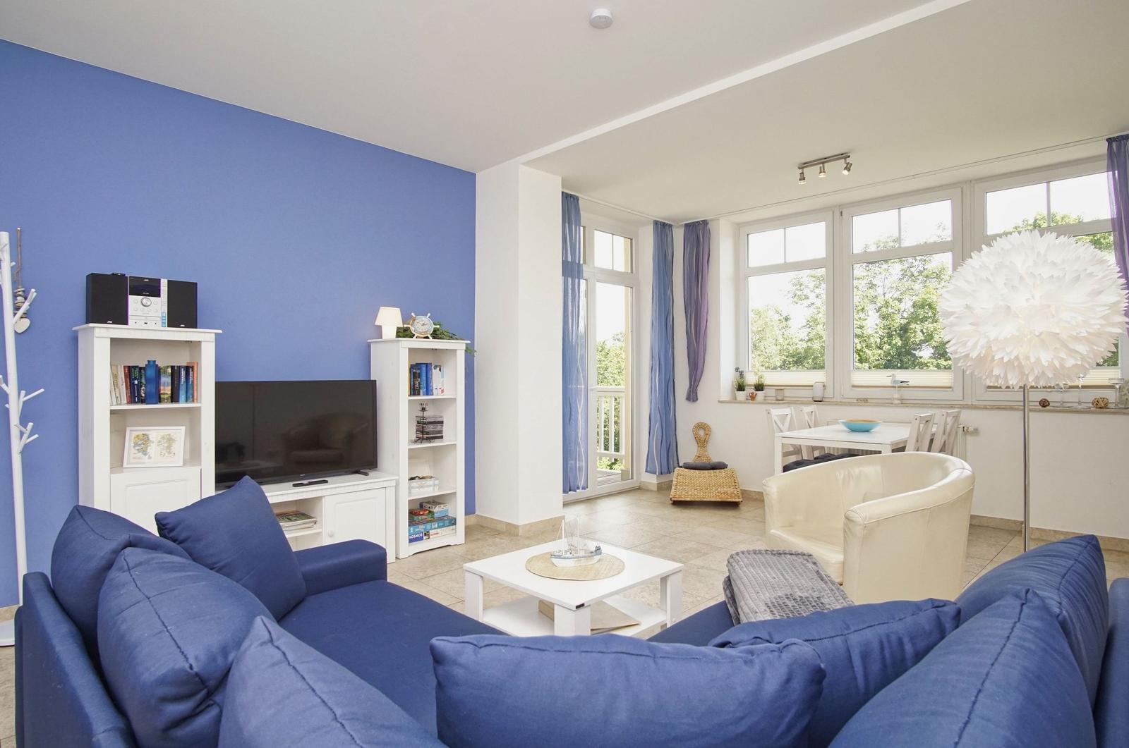 Wohnzimmer mit blauem Sofa, Fernseher und großem Fenster. Essbereich mit Tisch und Stühlen.