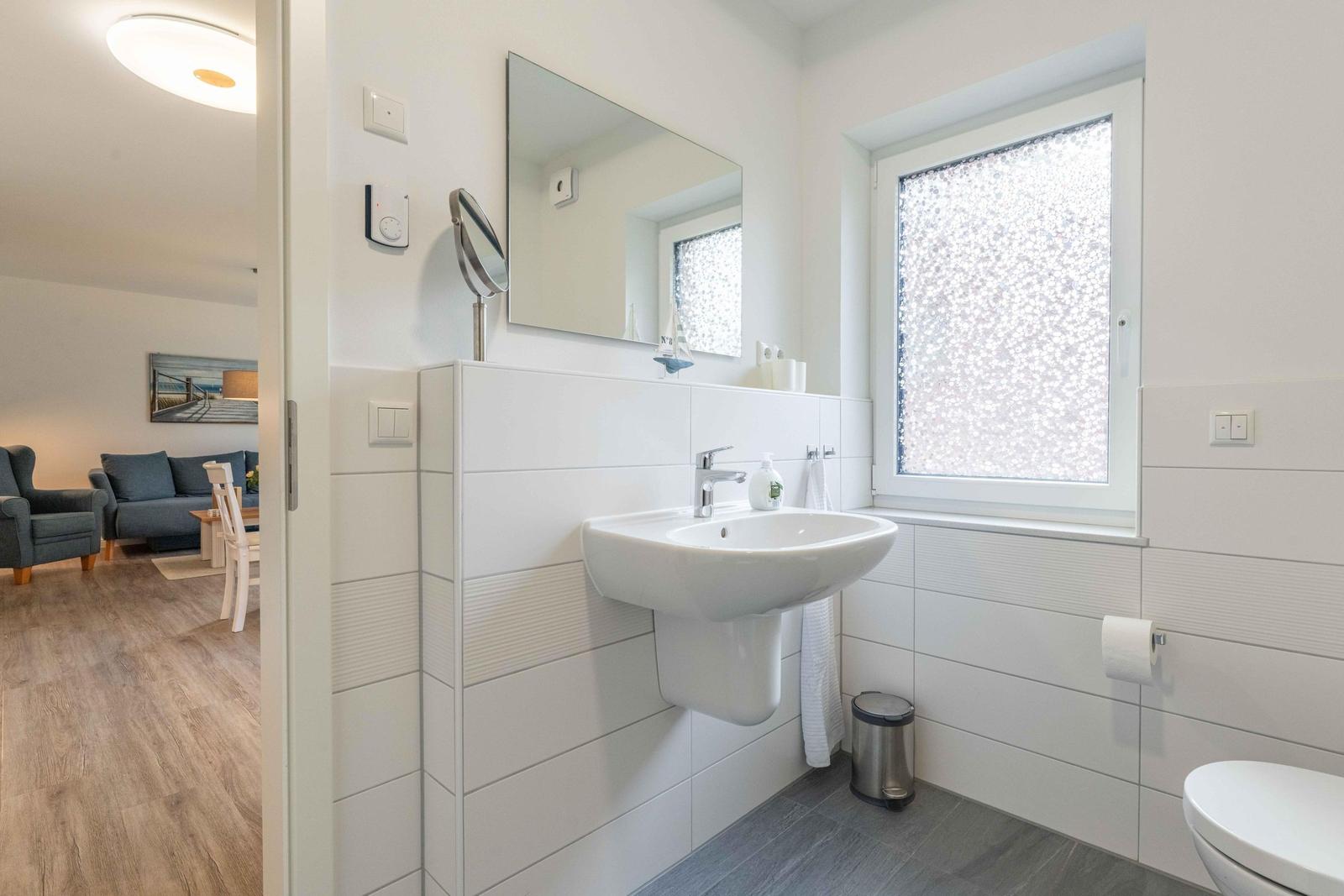 Badezimmer mit Waschbecken, Spiegel und Fenster. Im Hintergrund ist das Wohnzimmer sichtbar.