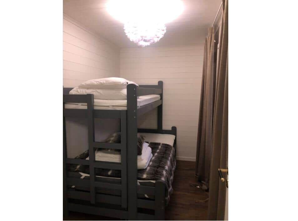 Zimmer mit Doppelstockbett, Holzboden und weißen Wänden. Deckenleuchte und Vorhang sichtbar.