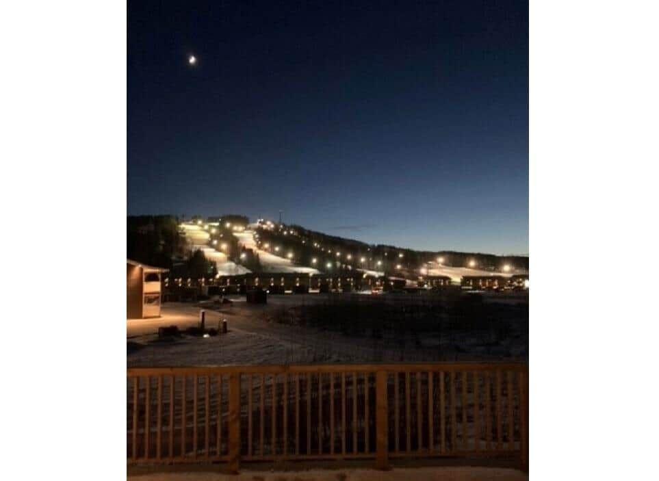 Nachtansicht eines beleuchteten Skigebiets mit Mond am Himmel und Holzgeländer im Vordergrund.