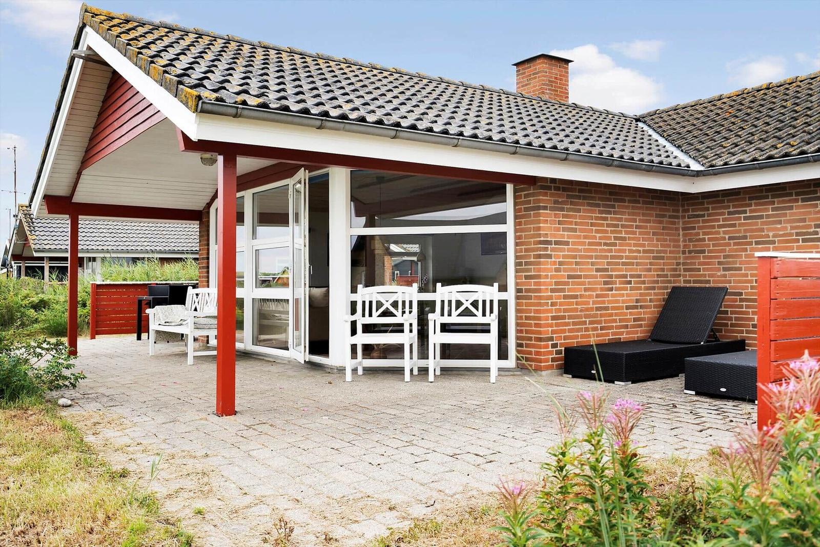 Haus mit Terrasse, weißen Stühlen und schwarzen Liegen. Roter Säulen und Ziegelmauer.