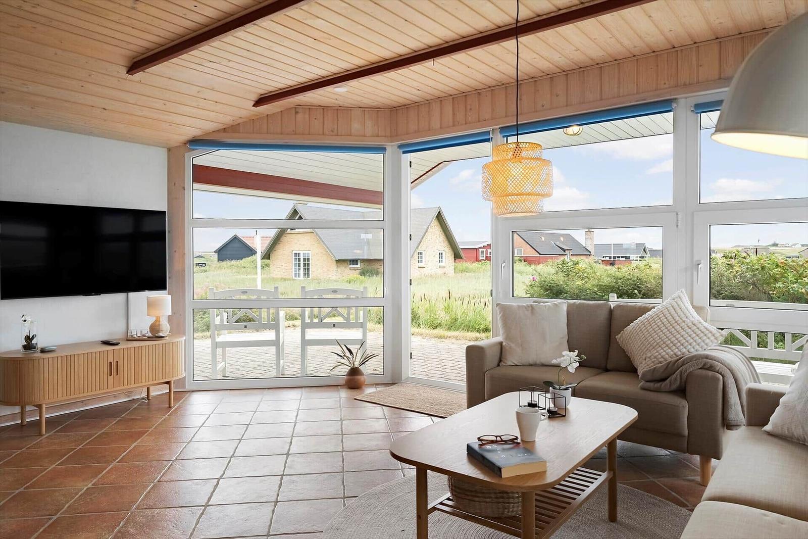 Wohnbereich mit Sofa, Couchtisch und Fernseher. Durchgehende Glasfront mit Blick auf Garten und Häuser.