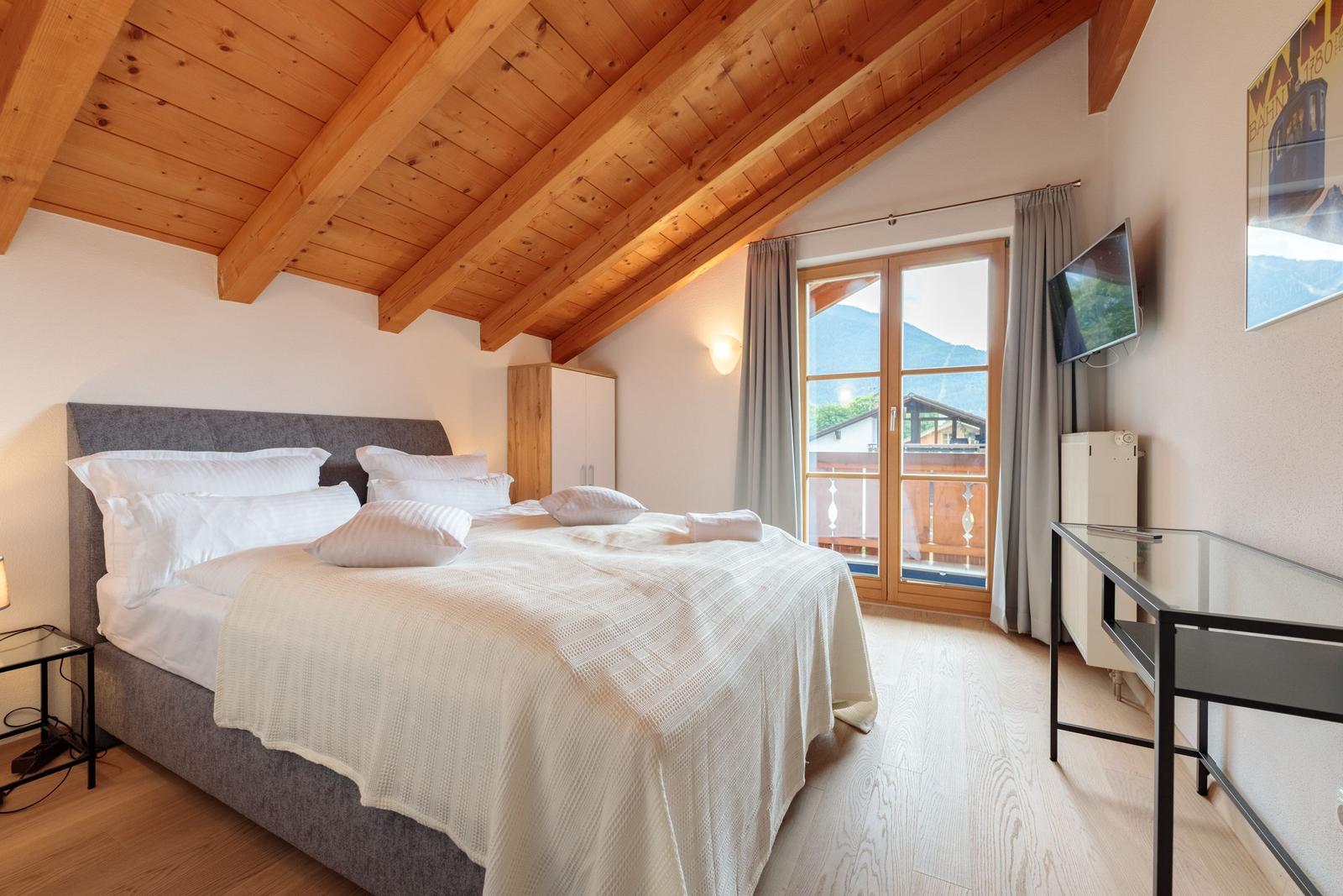 Schlafzimmer mit Balkon, Holzdecke und TV. Bett mit weißen Kissen.