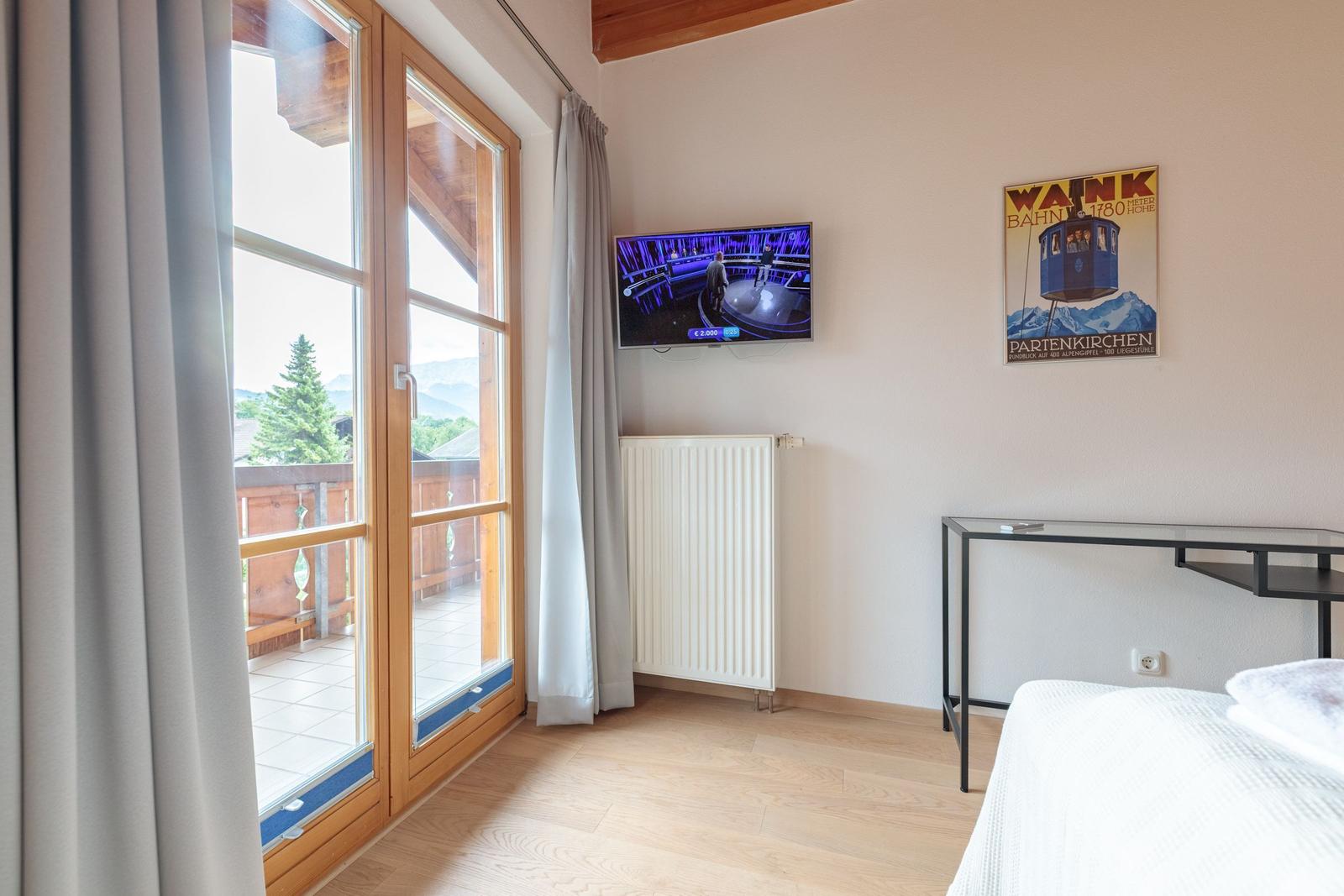 Zimmer mit TV, Balkon und Wandposter. Holzfußboden und weißer Heizkörper.