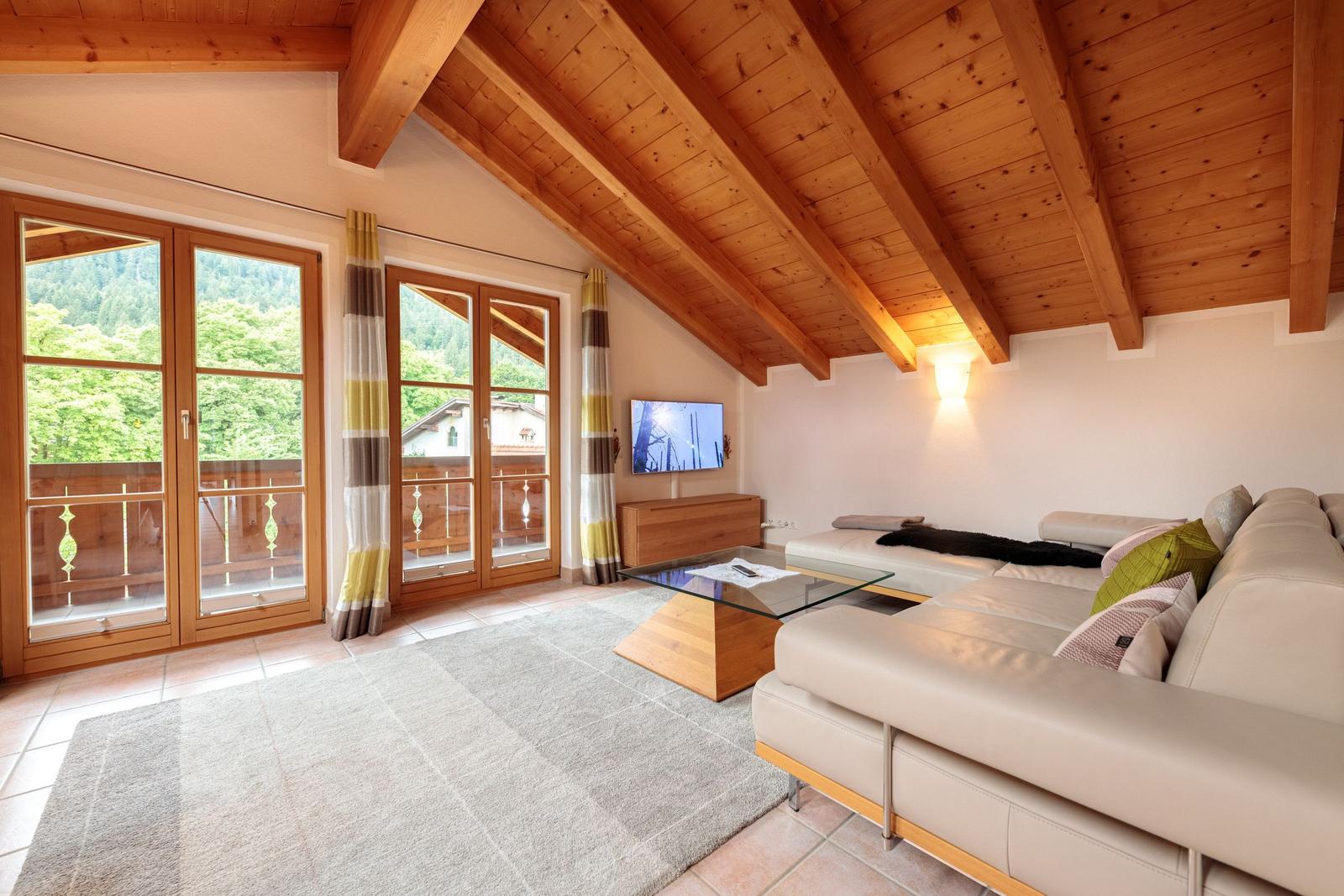 Wohnzimmer mit Holzdecke, Sofa, Fernseher und Blick auf Balkon und Wald.