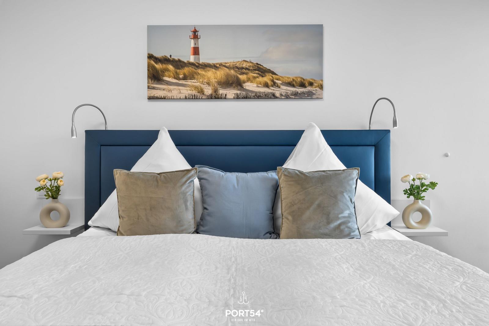 Schlafzimmer mit blauem Kopfteil, weißen Kissen und Wandbild mit Leuchtturm.