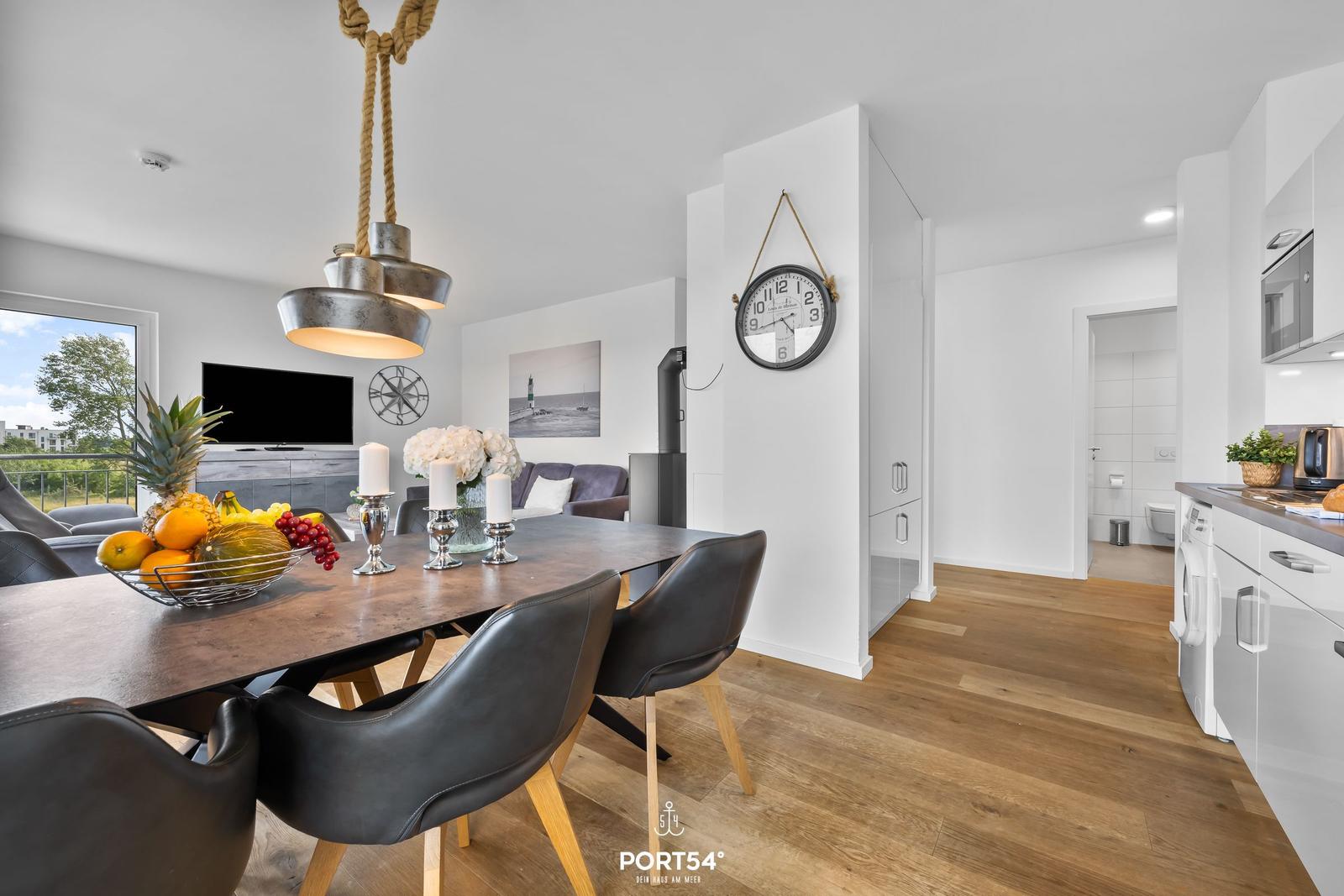 Essbereich mit Tisch, Stühlen und Obst. Küche, Wohnzimmer und Bad sind sichtbar.