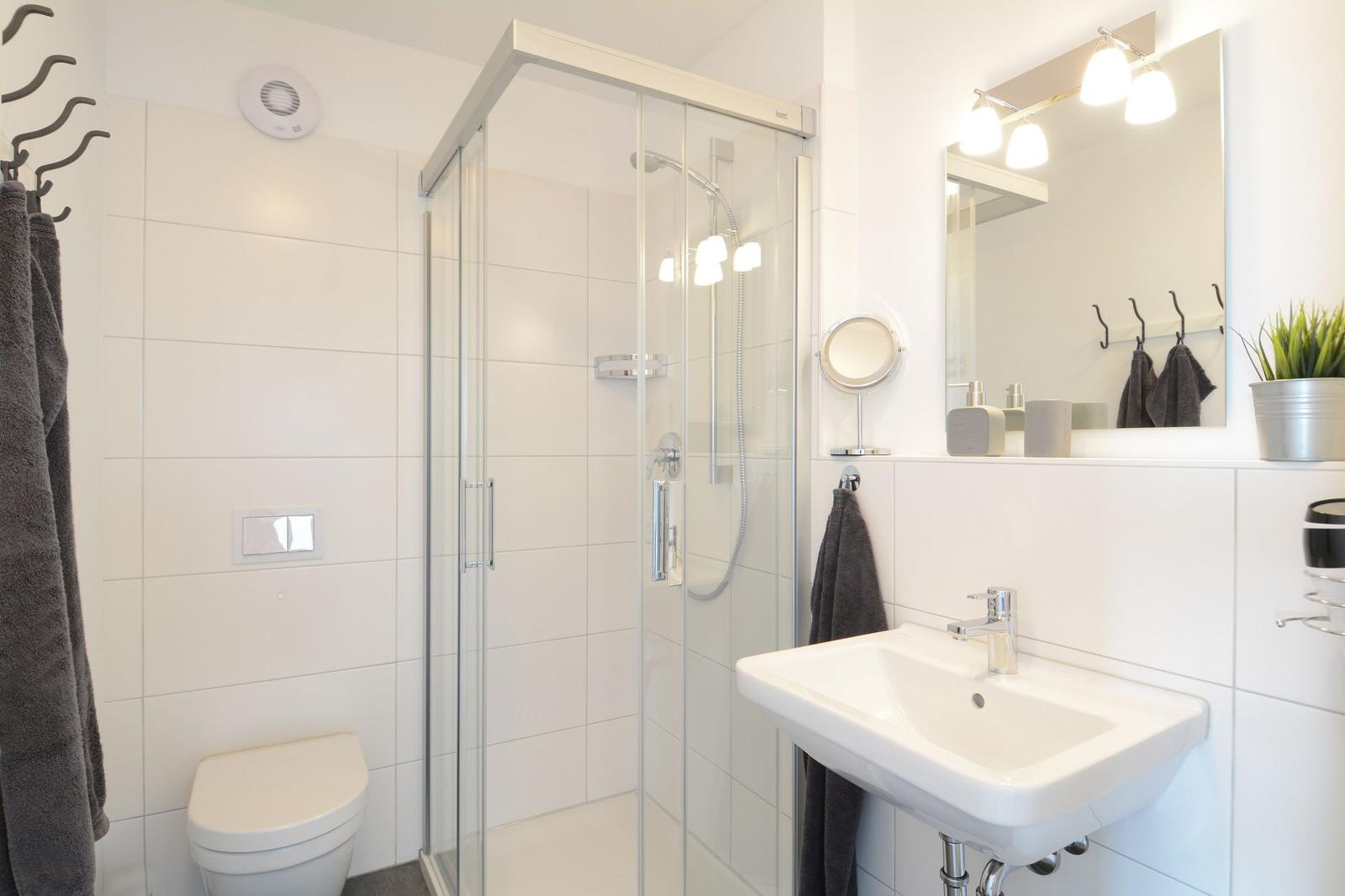 Badezimmer mit Dusche, Waschbecken und Toilette. Weiße Fliesen und graue Handtücher.